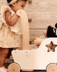 Ali + Oli Muslin Cotton Baby Bib in Ivory placed next to a doll in a wooden toy wagon