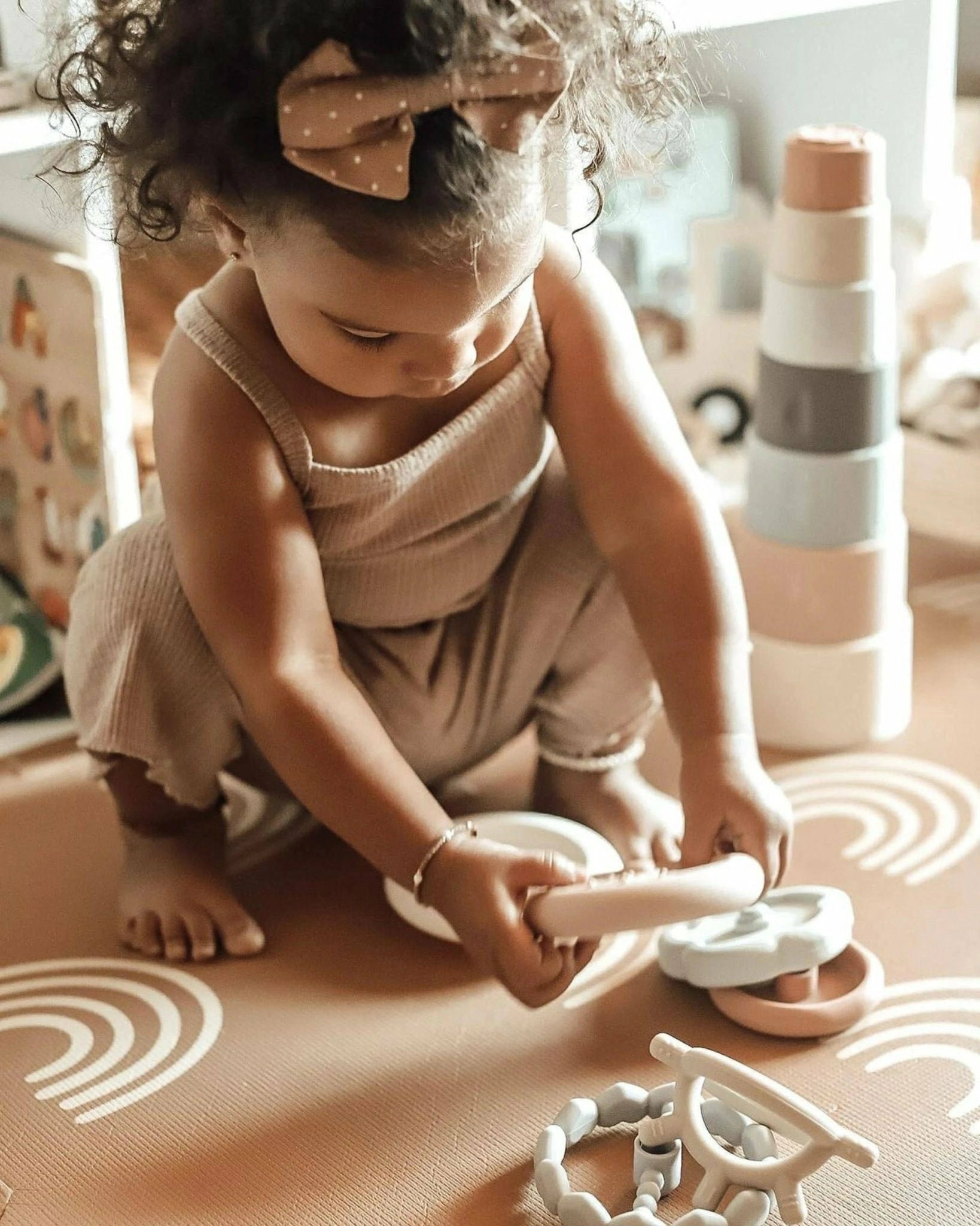 A toddler girl playing with the Ali + Oli Soft Silicone Stacking Ring Tower on a patterned rug