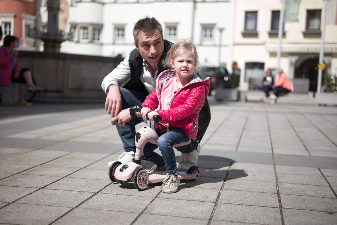 Toddler learning to balance on the rose Highwaykick 1 scooter with the assistance of an adult