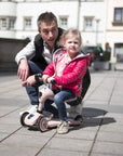 Toddler learning to balance on the rose Highwaykick 1 scooter with the assistance of an adult