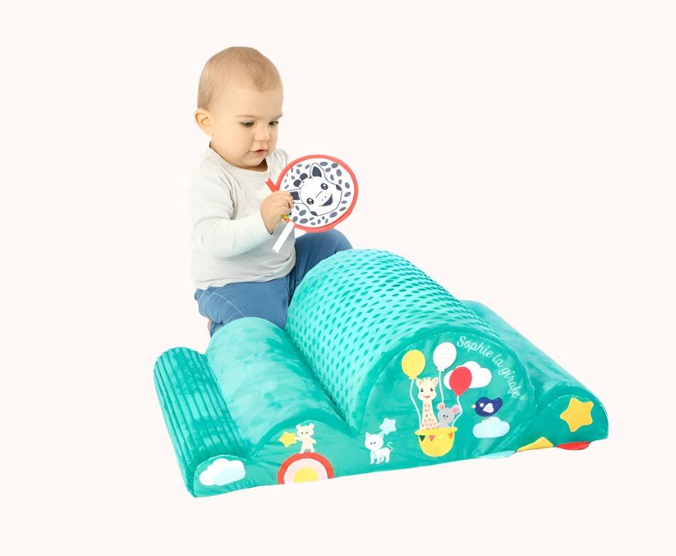 Toddler seated on a green Sophie La Girafe playmat with attached mirror toy